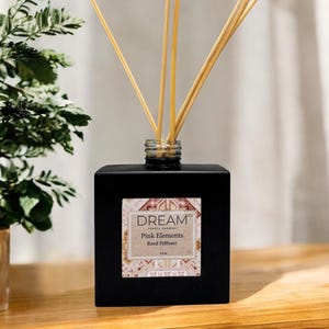 May include: A black square reed diffuser with the text "DREAM Pink Elements Reed Diffuser" on a label. The diffuser has several wooden reeds inserted into the top. A potted plant with green and white leaves is to the left.