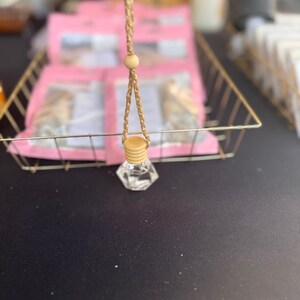 以下が含まれることがあります： 木製のキャップと、吊り下げ用の編み込みタンコードが付いた、透明なファセットガラスボトル。 ボトルは、ピンクのパッケージが入った金色のワイヤーバスケットの前に吊り下げられています。 背景はぼやけています。