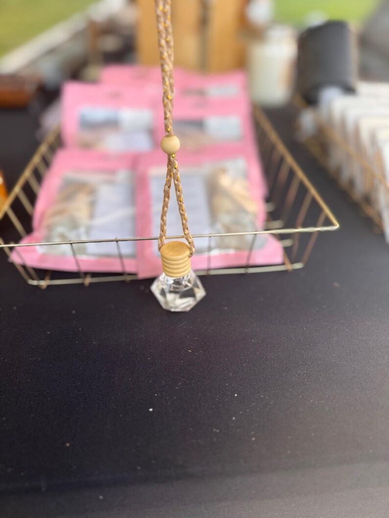 以下が含まれることがあります： 木製のキャップと、吊り下げ用の編み込みタンコードが付いた、透明なファセットガラスボトル。 ボトルは、ピンクのパッケージが入った金色のワイヤーバスケットの前に吊り下げられています。 背景はぼやけています。