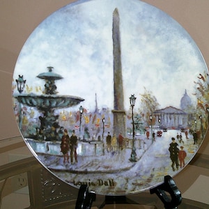 May include: A round ceramic plate with a painted image of a Parisian street scene. The scene features a fountain, an obelisk, and a building with a dome. The plate is signed "Louis Dall" in the lower right corner.