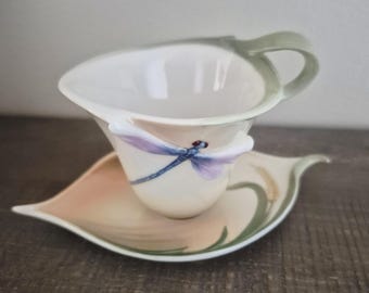 May include: A ceramic teacup and saucer set shaped like a leaf. The cup features a sculpted handle and a painted purple and blue dragonfly on the side. The saucer is cream-colored with soft pink shading and green leaf vein details along the edges.