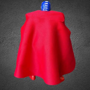 Puede incluir: Una peque&ntilde;a figura de acci&oacute;n que lleva una capa de tela roja vibrante que cae sobre la espalda. La figura tiene una cabeza azul con rayas verticales blancas y pies de pl&aacute;stico rojo visibles bajo el dobladillo de la capa.