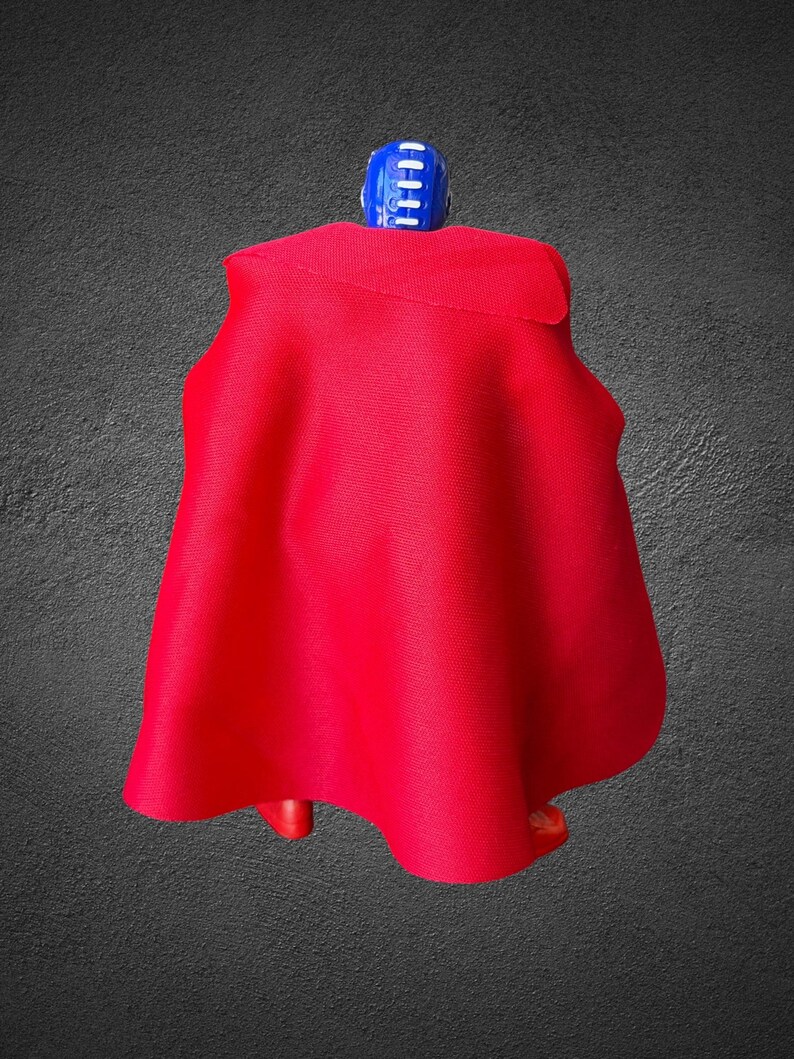 Puede incluir: Una peque&ntilde;a figura de acci&oacute;n que lleva una capa de tela roja vibrante que cae sobre la espalda. La figura tiene una cabeza azul con rayas verticales blancas y pies de pl&aacute;stico rojo visibles bajo el dobladillo de la capa.