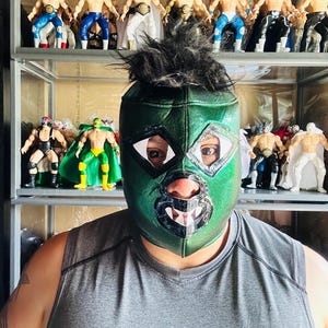 May include: A person wearing a green lucha libre mask with black hair. Behind the person are shelves filled with lucha libre action figures in various colors and outfits.