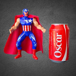 Puede incluir: Una figura de acci&oacute;n de pl&aacute;stico de un luchador enmascarado con un traje azul y rojo con una estrella y rayas blancas, junto a una capa roja, al lado de una lata de refresco roja con el nombre Oscar impreso en letras blancas en el lateral.