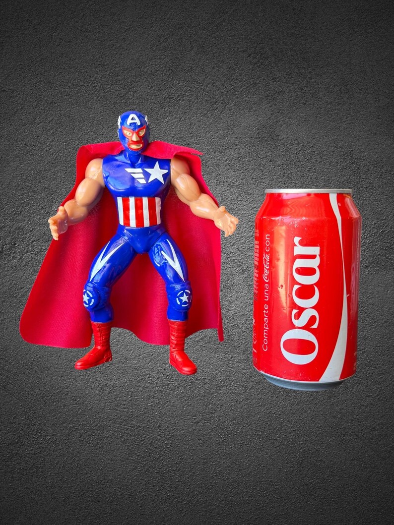Puede incluir: Una figura de acci&oacute;n de pl&aacute;stico de un luchador enmascarado con un traje azul y rojo con una estrella y rayas blancas, junto a una capa roja, al lado de una lata de refresco roja con el nombre Oscar impreso en letras blancas en el lateral.