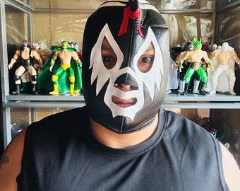 May include: A black and white lucha libre mask with a red accent, worn by a person. The mask features a stylized design with white eye and mouth openings. Action figures of wrestlers are displayed on shelves in the background.
