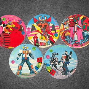 May include: Five vintage circular collectible cards featuring colorful illustrations of robots and mecha. Each card has a unique design with vibrant colors, including red, blue, and yellow. Some cards have Japanese text and numbers.