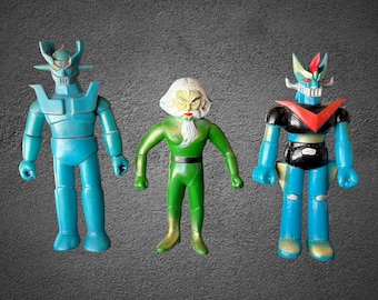 Puede incluir: Tres figuras de acción de pie. La figura de la izquierda es de color verde azulado sólido con un casco con cuernos. La figura central es un personaje vestido de verde con cabello y barba blancos. La figura de la derecha es azul y negra.
