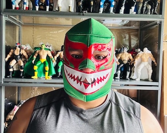 May include: A person wearing a green and red lucha libre mask with a white jagged-tooth design. The person is wearing a gray tank top. Shelves behind the person display multiple action figures of wrestlers in various outfits.