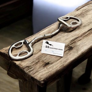Può includere: Un morso da cavallo argentato lucido con un design curvo, appoggiato su una superficie di legno rustico. Il morso presenta due anelli a D e una barra curva di collegamento. È presente anche un biglietto da visita con il testo "SS Equestrian Gears".