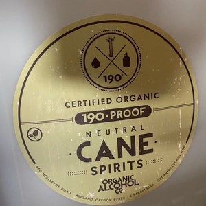 Puede incluir: Una etiqueta circular dorada y marr&oacute;n con el texto "Certified Organic 190-Proof Neutral Cane Spirits Organic Alcohol Co. 650 Mistletoe Road Ashland, Oregon 97520 (541) 201-1050 OrganicAlcohol.com" y un logotipo de un tallo de ca&ntilde;a de az&uacute;car, una gota de l&iacute;quido y una botella con el n&uacute;mero 190 grados.