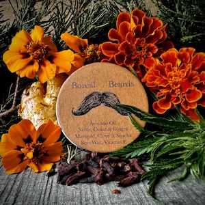 Könnte beinhalten: Ein runder, brauner Behälter mit dem Text "Boreal Beards" und einer Schnurrbart-Grafik. Der Behälter ist von orangefarbenen Ringelblumen, grünem Laub und Nelken umgeben. Die aufgeführten Zutaten sind Avocadoöl, Brennnessel, Zeder & Ingwer, Ringelblume, Nelke & Sencha, Bienenwachs und Vitamin E.