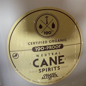 May include: A white plastic container with a yellow and brown circular label. The label says "Certified Organic 190 Proof Neutral Cane Spirits" and includes a logo with a sugarcane stalk, a drop of liquid, and a bottle. The label also includes the company name "Organic Alcohol Co.", address, and phone number.