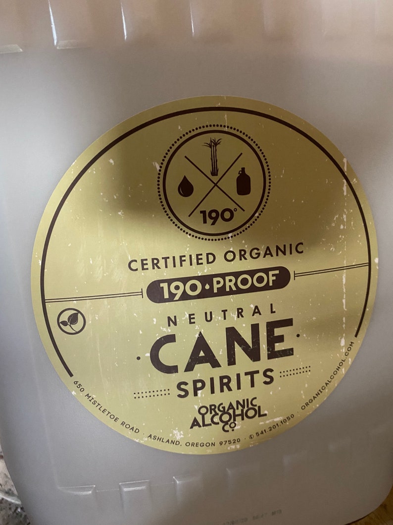 May include: A white plastic container with a yellow and brown circular label. The label says "Certified Organic 190 Proof Neutral Cane Spirits" and includes a logo with a sugarcane stalk, a drop of liquid, and a bottle. The label also includes the company name "Organic Alcohol Co.", address, and phone number.