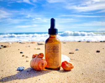 Puede incluir: Una botella de vidrio marrón de extracto de raíz de cúrcuma Boreal Medicine con un cuentagotas negro, colocada en una playa arenosa con raíces de cúrcuma. La etiqueta incluye el texto "Turmeric Root Curcuma Longa". El fondo muestra el océano y el cielo azul.