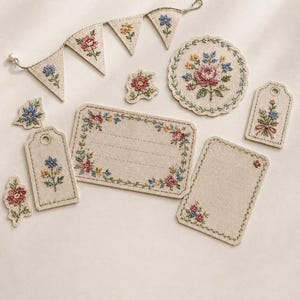 May include: A collection of beige linen fabric items featuring delicate floral embroidery in pink, blue, and yellow. The set includes a triangular bunting banner, a circular scalloped coaster, two rectangular gift tags with eyelets, and two blank labels.