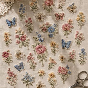 May include: A collection of embroidered fabric patches featuring various floral arrangements, butterflies, and bees in shades of pink, blue, yellow, and green. The patches are arranged on a light beige fabric surface next to a pair of metal scissors.