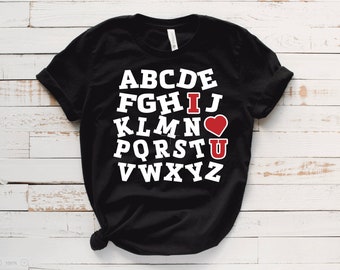 May include: Black t-shirt with a white alphabet design. The letters "I" and "U" are highlighted in red, with a red heart symbol between them. The shirt is displayed on a white wooden surface.