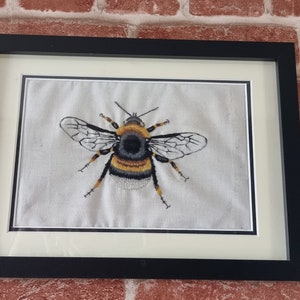 May include: A framed embroidery of a bumblebee with black and yellow stripes, white wings, and a black head. The embroidery is on a white fabric and is framed in a black frame.