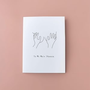 May include: A white card featuring a minimalist line drawing of two hands reaching out. The text "To My Main Squeeze" is printed beneath the illustration. The background is a solid, muted pink. A simple, romantic design.