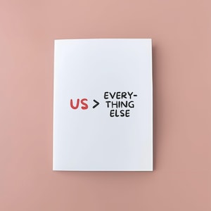 May include: A white card with the words "US > EVERYTHING ELSE" in black and red lettering. The card is centered on a pink background.
