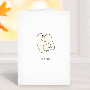 May include: A white greeting card with a minimalist illustration of two hugging ghosts. The text "HEY BOO" is printed in black below the ghosts. The card is set against a blurred background with fall foliage.