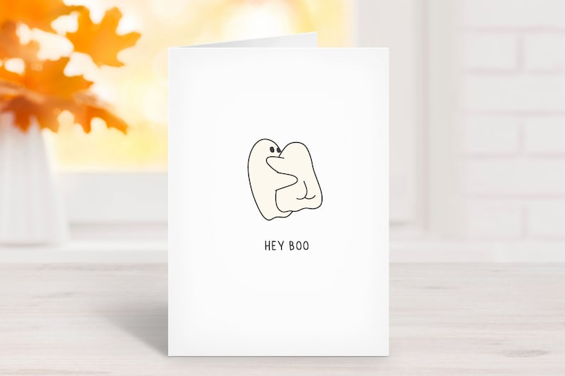 May include: A white greeting card with a minimalist illustration of two hugging ghosts. The text "HEY BOO" is printed in black below the ghosts. The card is set against a blurred background with fall foliage.