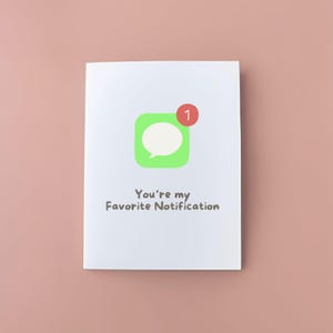 Puede incluir: Una tarjeta de felicitación blanca con un icono de bocadillo verde y una insignia de notificación roja. El texto "You're my Favorite Notification" está impreso debajo. El fondo es de color rosa liso.