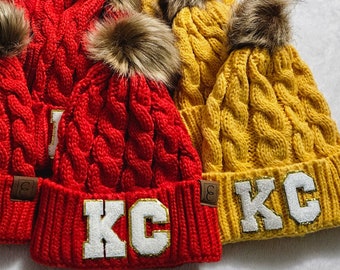 May include: Red and yellow cable knit beanies with faux fur pom-poms. Each beanie features the letters "KC" in white with gold trim. The beanies are displayed on a white surface.