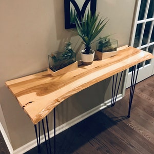May include: A light wood console table with a live edge top and black hairpin legs. The table is decorated with two small potted plants and a larger potted plant.