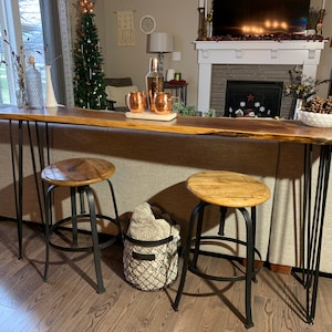 May include: A long, live edge wood table with black hairpin legs. Two black metal bar stools with wood seats are placed in front of the table. The table is decorated with a clear glass bottle, two copper mugs, and a white bowl with decorative white stones.