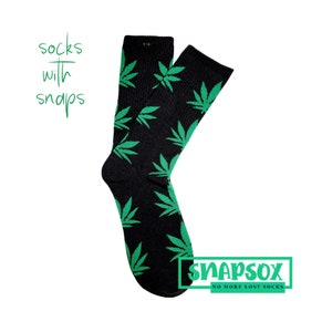 Könnte beinhalten: Schwarze Socken mit einem grünen Marihuana-Blatt-Muster. Die Socken haben einen Druckknopfverschluss oben. Der Text "Snapsox No More Lost Socks" ist unten im Bild gedruckt.