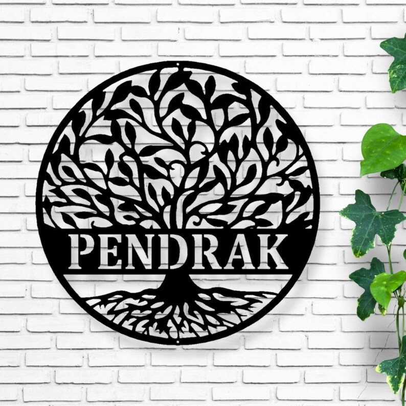 May include: Black metal tree of life wall art with the name PENDRAK cut out in a circular design. The tree has detailed leaves and roots. The art is mounted on a white brick wall.