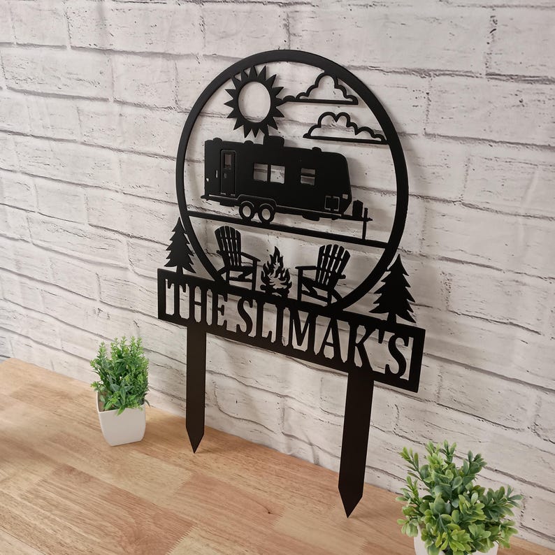 May include: Black metal sign featuring a camper, sun, clouds, trees, chairs, and a campfire. The sign reads "THE SLIMAR'S" in bold, capital letters. The sign is designed for outdoor use and is a decorative item for a campsite or RV.
