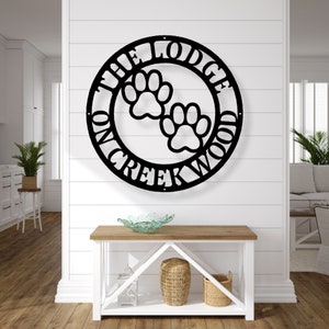 May include: Black metal wall art featuring a circular design with the text "THE LODGE ON CREEKWOOD" and two paw prints. The sign is mounted on a white wall above a wooden console table with decorative items.
