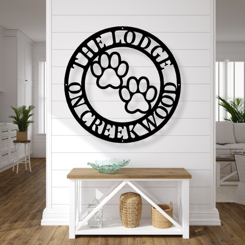 May include: Black metal wall art featuring a circular design with the text "THE LODGE ON CREEKWOOD" and two paw prints. The sign is mounted on a white wall above a wooden console table with decorative items.