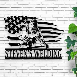May include: Black metal silhouette of a welder with a welding mask and a welding torch, in front of an American flag, with the text "STEVEN'S WELDING" below.