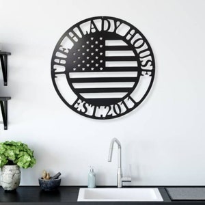 K&ouml;nnte beinhalten: Schwarze Metall-Wandkunst in Kreisform mit dem Design der amerikanischen Flagge. Der Text "THE LADY HOUSE EST. 2011" ist um die Flagge herum ausgeschnitten. An einer wei&szlig;en Wand &uuml;ber einem K&uuml;chenbecken angebracht.