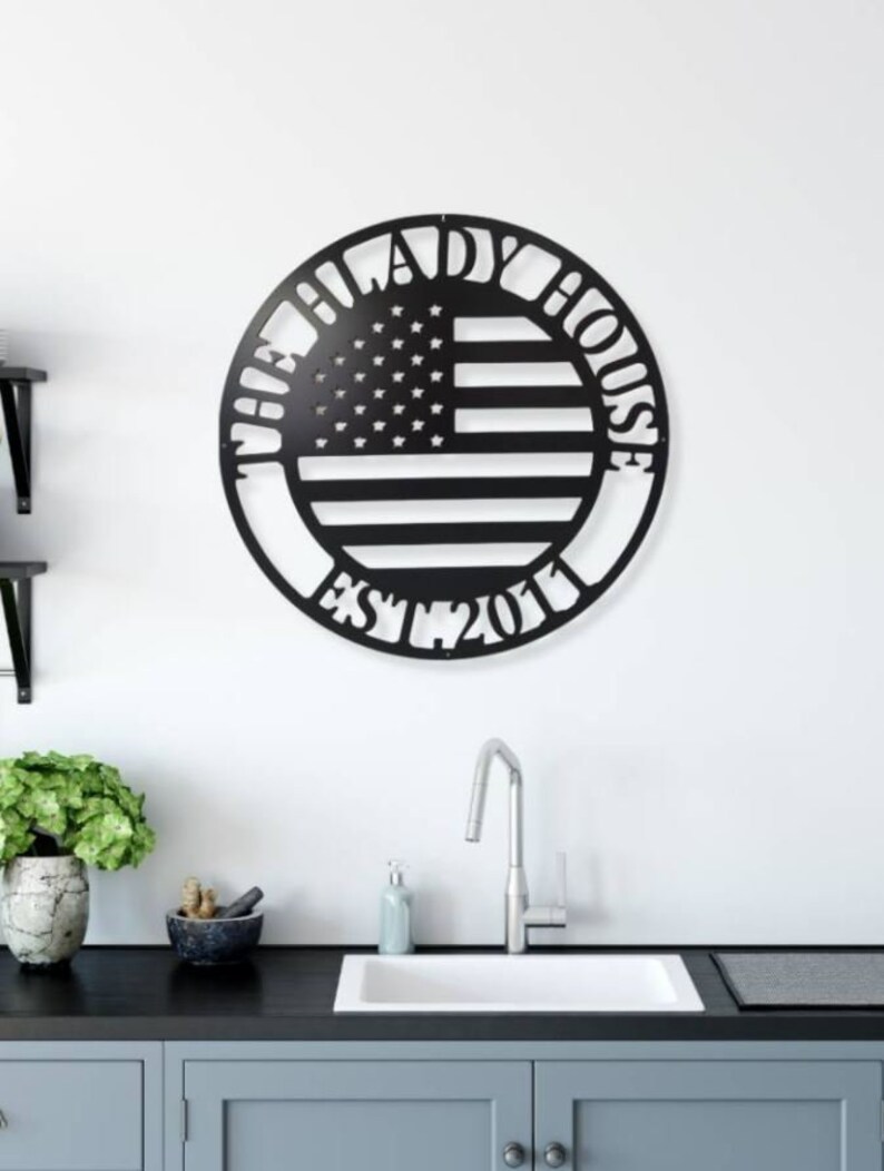 K&ouml;nnte beinhalten: Schwarze Metall-Wandkunst in Kreisform mit dem Design der amerikanischen Flagge. Der Text "THE LADY HOUSE EST. 2011" ist um die Flagge herum ausgeschnitten. An einer wei&szlig;en Wand &uuml;ber einem K&uuml;chenbecken angebracht.