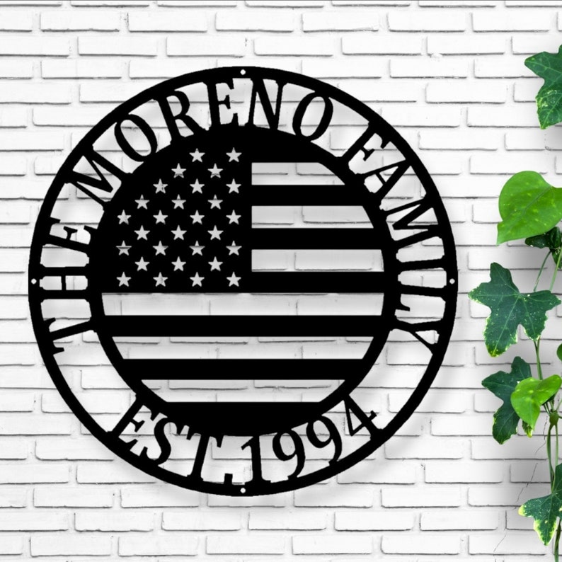 K&ouml;nnte beinhalten: Ein schwarzes Metallschild mit dem Design der amerikanischen Flagge. Das runde Schild hat den Text "THE MORENO FAMILY" oben und "EST. 1994" unten. Das Flaggen-Design befindet sich in der Mitte.
