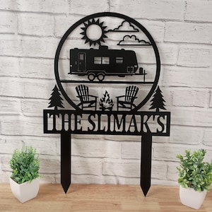May include: Black metal camping-themed sign featuring a camper, sun, clouds, two chairs, a campfire, and two trees. The sign includes the text "THE SLIMAK'S" and is designed to be placed in the ground. Perfect for outdoor decor.