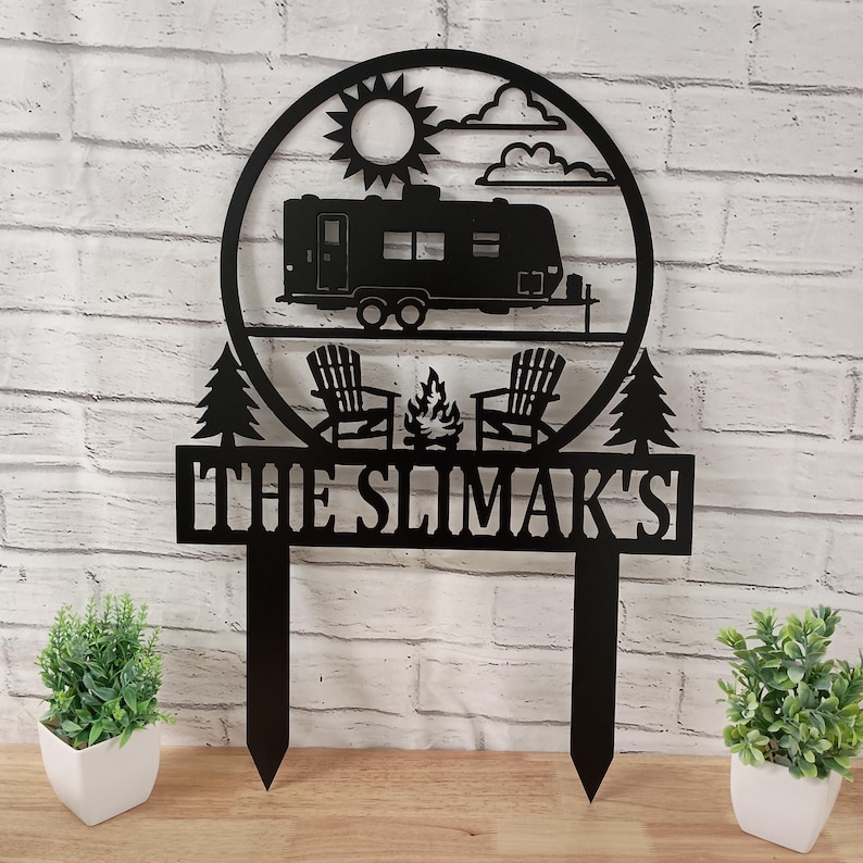 May include: Black metal camping-themed sign featuring a camper, sun, clouds, two chairs, a campfire, and two trees. The sign includes the text "THE SLIMAK'S" and is designed to be placed in the ground. Perfect for outdoor decor.