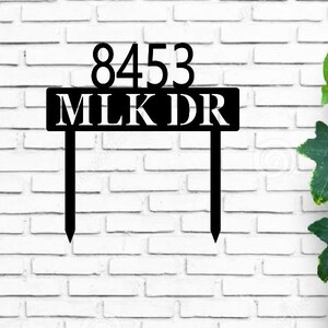 May include: Black metal house number sign with the numbers 8453 and the street name MLK DR. The sign is attached to two metal stakes.