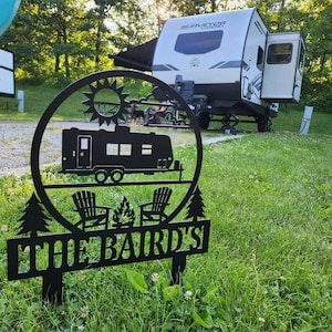 May include: Black metal yard sign featuring a camping scene with a trailer, chairs, and a campfire. The sign is circular with a sun design at the top and the text "THE BAIRD'S" at the bottom. A white and gray camper is in the background.