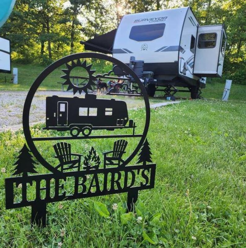May include: Black metal yard sign featuring a camping scene with a trailer, chairs, and a campfire. The sign is circular with a sun design at the top and the text "THE BAIRD'S" at the bottom. A white and gray camper is in the background.