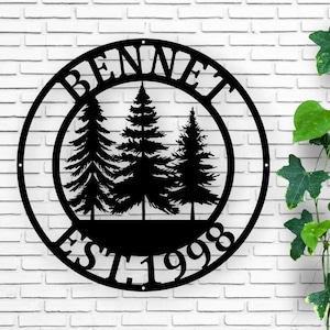May include: Black metal wall art featuring the name "BENNET" in a circular design. Inside the circle are three stylized pine trees and the text "EST 1998". The art is mounted on a white brick wall.