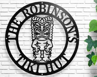 May include: Black metal wall art depicting a Tiki figure inside a circular frame. The words "THE ROBINSONS" and "TIKI HUT" are cut out around the frame. It's mounted on a white brick wall.