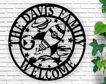 May include: Black metal family welcome sign with a space theme. The circular sign features a rocket ship, planets, stars, and the words "THE DAVIS FAMILY WELCOME" cut out in a decorative font. The sign is mounted on a white brick wall.