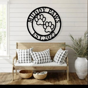 May include: Black metal wall art with the words "Muddy Paws" and "EST. 2022" surrounding two paw prints. The sign is displayed above a wooden bench with checkered pillows and woven baskets.